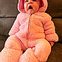 baby, child, pink_onesie, fluffy_clothing, hooded_jacket, sitting, couch, indoor, expression, surprised, cute, soft_texture, warm_clothing, portrait, infant, face, hand, footwear, cozy, casual