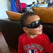 Leo joined the competition — help win amazing prizes! toddler, child, sunglasses, red_shirt, indoor, smile, curly_hair, couch, pillow, wood_floor, toy, casual_clothing, person, portrait, happy, young_child, fashion, living_room, sitting, cheerful