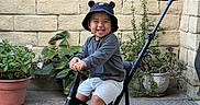 Leo joined the competition — help win amazing prizes! child, toddler, tricycle, hat, smiling, outdoor, plants, potted_plants, brick_wall, concrete_floor, shorts, long_sleeve_shirt, footwear, happy, toy_vehicle, handle, greenery, sunlight, casual_clothing, portrait