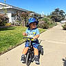 Leo is registered to the contest to win money with this photo: toddler, child, tricycle, blue_hat, blue_shirt, blue_shorts, sneakers, sidewalk, sunny_day, outdoor, residential_area, grass, bushes, trees, house, dinosaur_print, shadow, curious_expression, play, summer_clothing