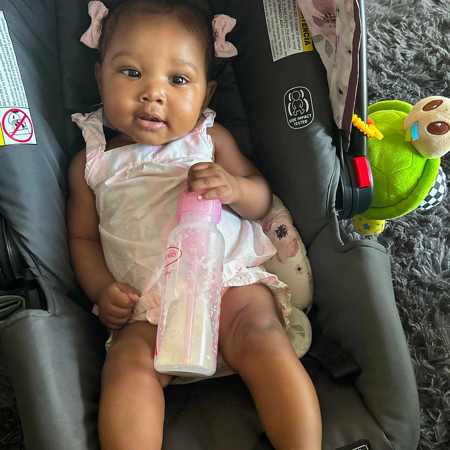 Noemi is registered to the contest to win money with this photo: baby, car_seat, child, cute, floor, footwear, hair_bows, happy, indoor, infant, milk_bottle, person, pink_dress, playful, rug, sandals, seat, smiling, toy, turtle_toy