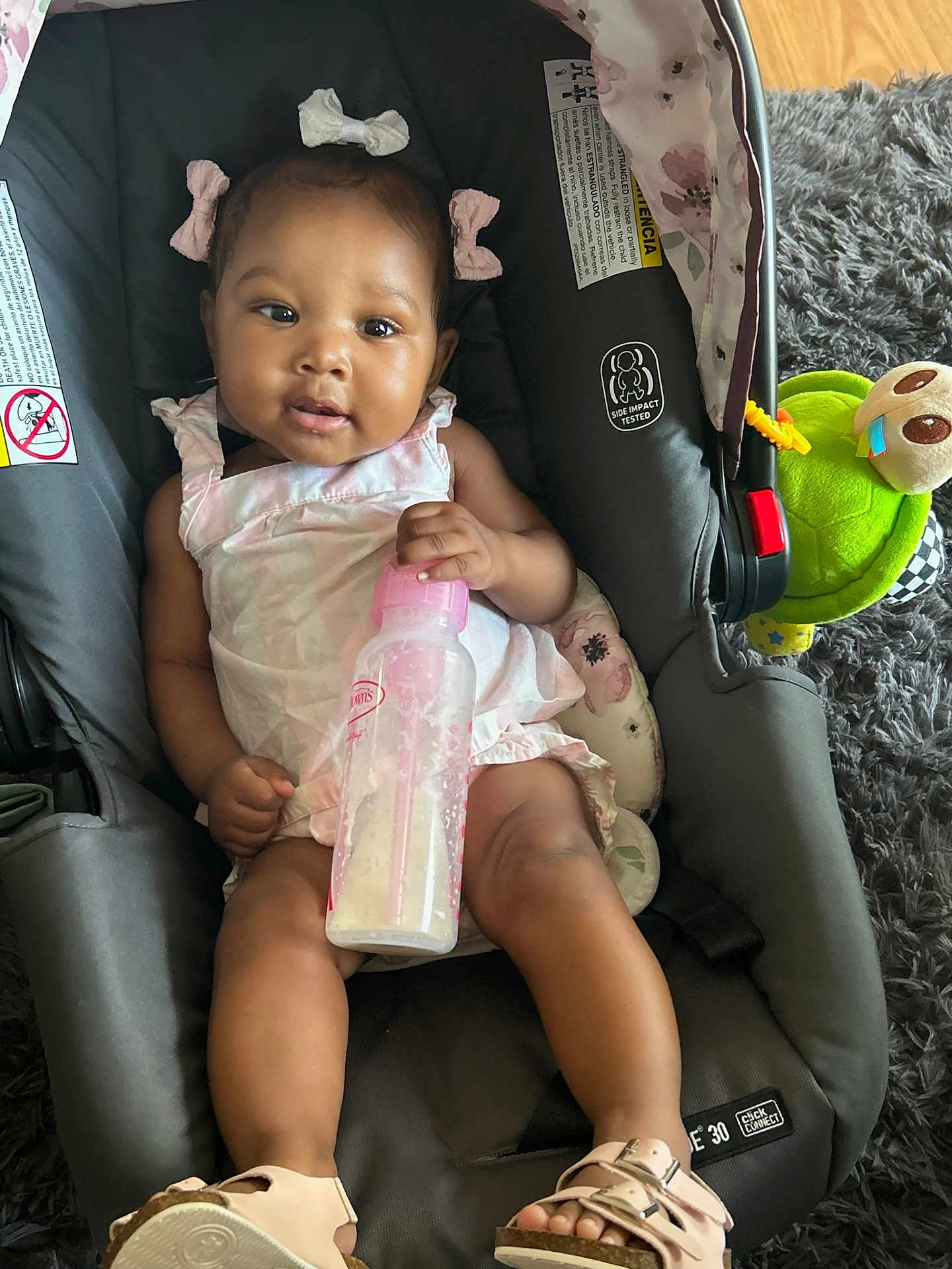 Noemi is registered to the contest to win money with this photo: baby, child, car_seat, milk_bottle, toy, turtle_toy, hair_bows, pink_dress, footwear, sandals, floor, rug, indoor, cute, smiling, infant, person, seat, happy, playful