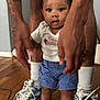 toddler, child, baby, red_bow, shorts, white_shirt, red_sandals, socks, sneakers, tattoo, legs, floor, indoor, person, hands, wooden_floor, curious, standing, casual_clothing, portrait