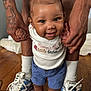 baby, child, red_bow, smiling, holding_hands, blue_shorts, white_shirt, sandals, adult_hands, tattoos, sneakers, wooden_floor, indoor, person, cute, happy, standing, support, family, closeup
