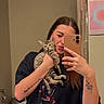 woman, cat, kitten, mirror, selfie, tattoo, bathroom, toilet, curtain, smartphone, black_tshirt, tabby_cat, young_adult, pouting, holding, indoor, reflection, casual, furniture, wooden_stool
