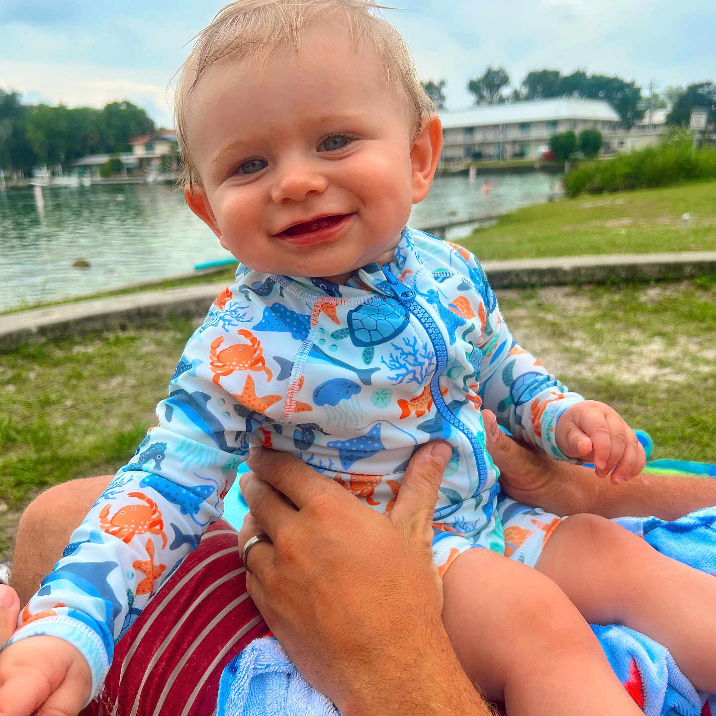 Brooks is registered to the contest to win money with this photo: baby, child, smile, swimsuit, ocean_theme, water, lake, outdoor, grass, adult_hand, person, nature, cloudy_sky, greenery, portrait, happy, summer, vacation, holding, playful
