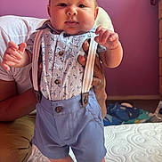 Konrad is registered to the contest to win money with this photo: baby, child, person, suspenders, blue_shorts, floral_shirt, indoor, bed, purple_wall, hand, smiling, cute, feet, portrait, infant, clothing, furniture, adult_hand, happy, standing