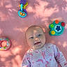 Octavia is registered to the contest to win money with this photo: baby, infant, smile, pink_blanket, toy, colorful_toy, playtime, child, dappled_light, happy, cute, portrait, soft_texture, indoors, face, clothing, onesie, lying_down, blue_eyes, shadows