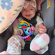 Ainsley is registered to the contest to win money with this photo: baby, smiling, car_seat, toy, blanket, headband, clothing, infant, happy, cute, child, seatbelt, pink, floral, indoors, portrait, person, footwear, bottle, colorful