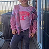 child, girl, smile, standing, pink_clothing, jacket, tshirt, leggings, sneakers, deck, railing, outdoor, daylight, hair, face, window, reflection, air_conditioner, pose, happy
