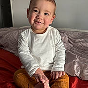 Yanis a rejoint le concours — aidez-le/la à gagner de superbes lots ! child, toddler, smile, bedroom, bed, blanket, white_shirt, orange_pants, toes, hands, portrait, sitting, cozy, natural_light, neutral_wall, rumpled_bedding, happy, cute, indoor, infant