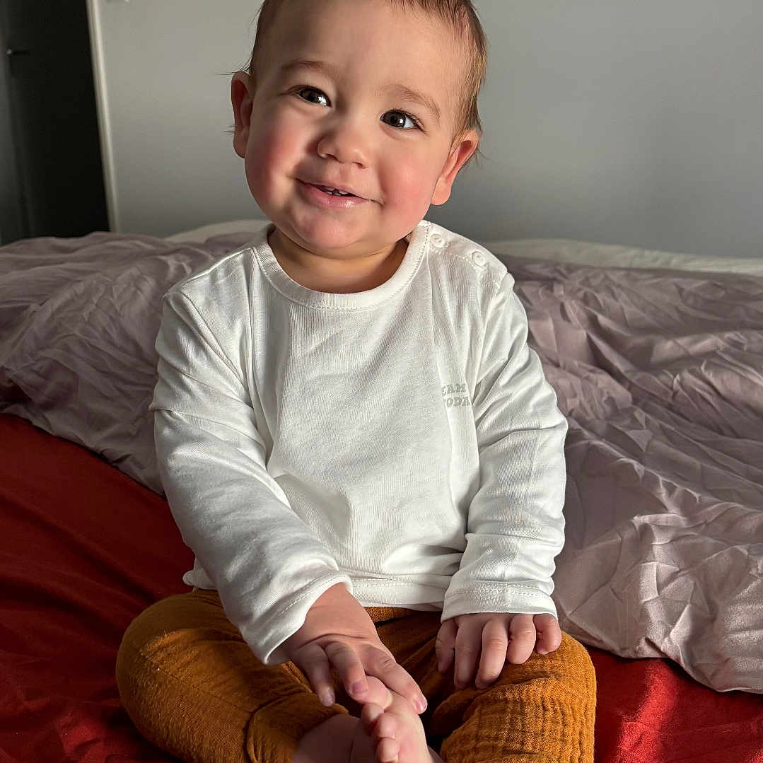 Yanis a rejoint le concours — aidez-le/la à gagner de superbes lots ! bed, bedroom, blanket, child, cozy, cute, hands, happy, indoor, infant, natural_light, neutral_wall, orange_pants, portrait, rumpled_bedding, sitting, smile, toddler, toes, white_shirt