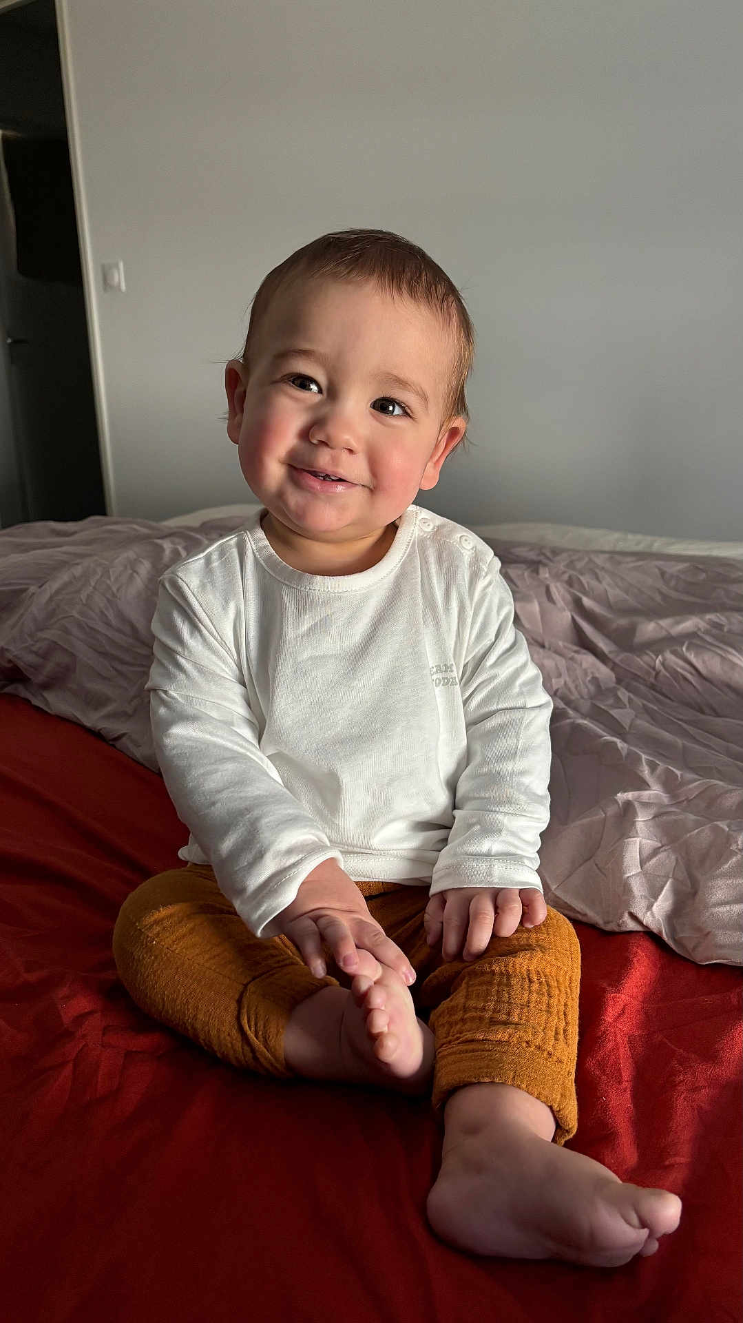 Yanis a rejoint le concours — aidez-le/la à gagner de superbes lots ! child, toddler, smile, bedroom, bed, blanket, white_shirt, orange_pants, toes, hands, portrait, sitting, cozy, natural_light, neutral_wall, rumpled_bedding, happy, cute, indoor, infant
