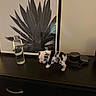 puppy, dog, black_and_white, water_bottle, headphones, table, drawer, artwork, plant_art, indoor, pet, animal, curious, small, spotted, wooden_furniture, neutral_wall, frame, decor, surface