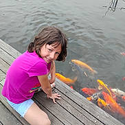 Natalee is registered to the contest to win money with this photo: child, girl, smiling, dock, water, koi_fish, fish, outdoor, summer, wood, casual_clothing, shorts, tshirt, nature, recreation, happy, playful, crouching, daytime, pond