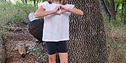 Madalynn is registered to the contest to win money with this photo: child, girl, outdoor, tree, forest, nature, smile, hand_gesture, heart_shape, white_shirt, black_shorts, socks, sandals, standing, grass, leaves, trunk, casual_clothing, young, happy