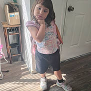 Kaytelinn is registered to the contest to win money with this photo: child, indoor, standing, pose, hand_on_chin, door, wooden_floor, cardboard_box, table, sunlight, shadow, shorts, shirt, sneakers, backpack, floor, wall, expression, hair, person
