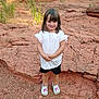 child, girl, smile, standing, rocky_terrain, outdoor, white_shirt, black_shorts, white_shoes, pink_design, grass, nature, rocks, young_child, casual_clothing, short_hair, daylight, happy, cute, portrait