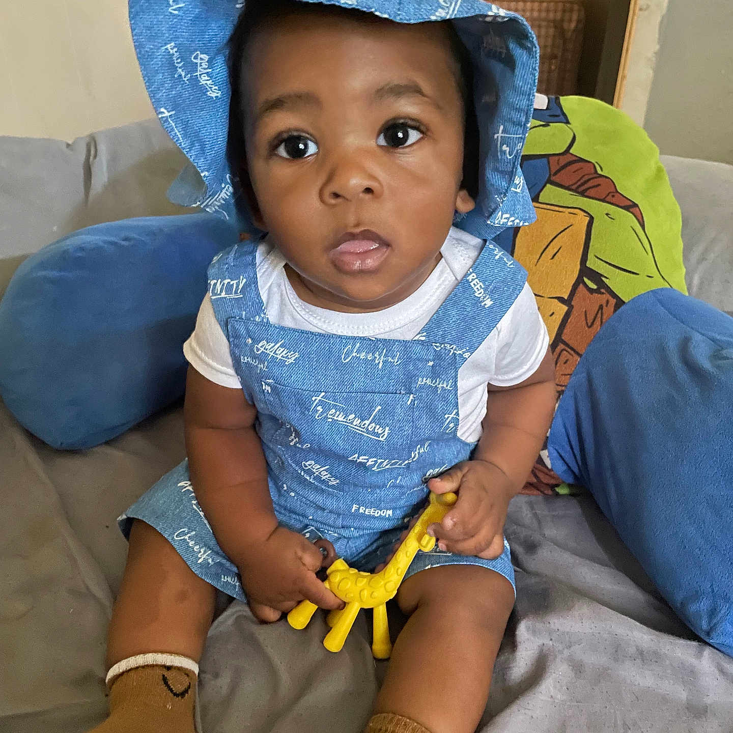 Khasan joined the competition — help win amazing prizes! baby, blanket, blue_hat, brown_skin, casual_clothing, child, couch, curious_expression, cushions, giraffe_toy, indoor, living_room, overalls, playful, seated, socks, toddler, toy, white_shirt, wide_eyes