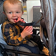 Reid is registered to the contest to win money with this photo: toddler, child, stroller, strap, yellow_checkered_shirt, black_checkered_shirt, hands, face, smile, indoor, bus_interior, shoes, jeans, seat, playful, hair, cute, person, clothing, transport