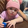 baby, child, toddler, pink_hat, pom_poms, purple_shirt, couch, leather, indoor, portrait, face, eyes, looking_at_camera, sitting, toy, blue_object, adult, tattoo, tile_floor, home
