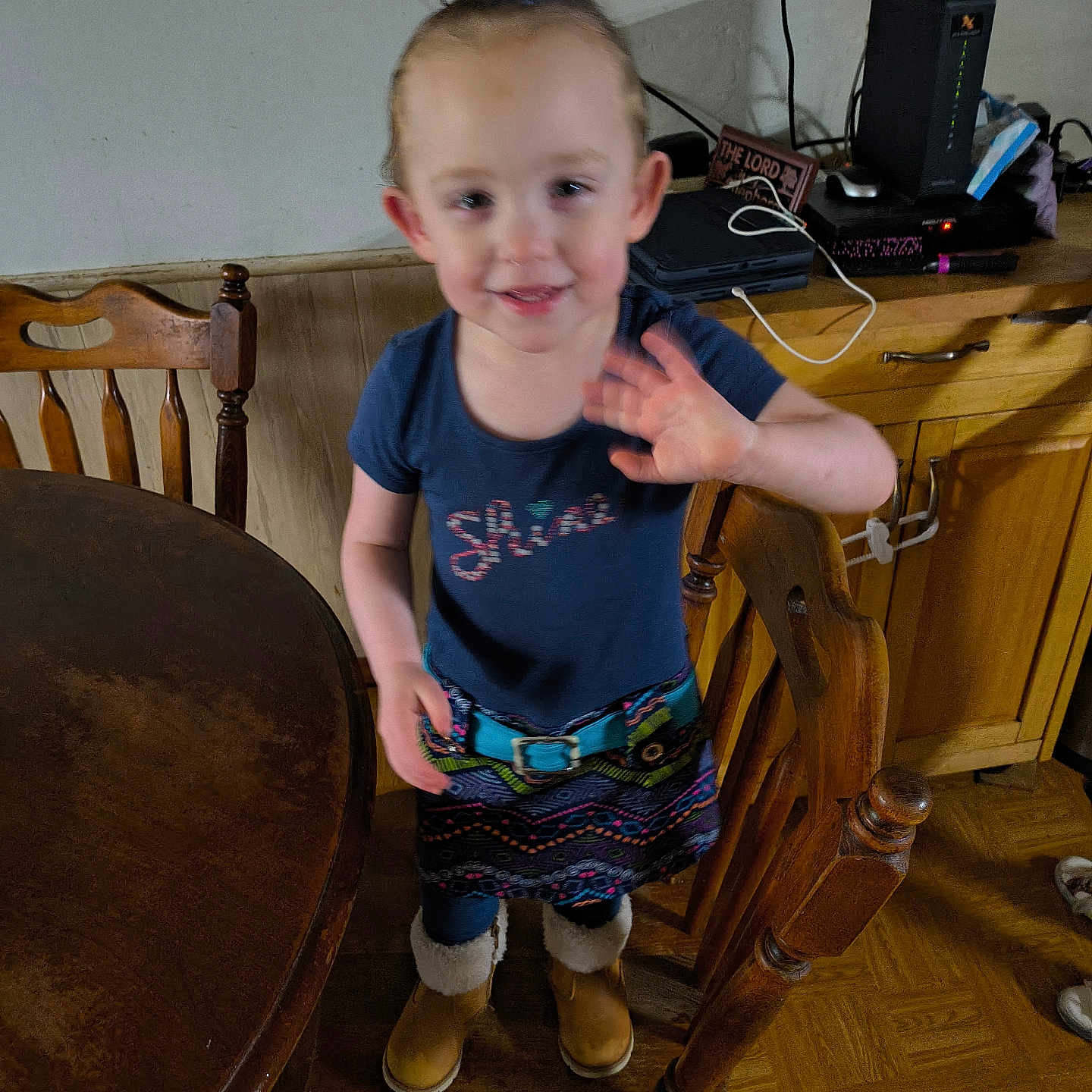 Scarlett is registered to the contest to win money with this photo: boots, cable, chair, child, face, floor, gadget, hair, hand, indoor, kitchen, person, shirt, skirt, smiling, standing, table, toddler, waving, wooden_furniture