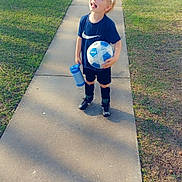 Gideon is registered to the contest to win money with this photo: child, boy, soccer_ball, water_bottle, sidewalk, grass, sunlight, visor, sportswear, shorts, socks, shoes, smiling, outdoor, daylight, recreation, happy, playing, leisure, neighborhood