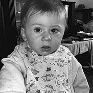 Liam participe au concours pour gagner de l'argent avec cette photo : toddler, child, indoor, bib, dinosaur_pattern, hand_holding, sitting, clothing, portrait, face, expression, hair, person, table, furniture, photograph, black_and_white, nail_polish, room, background