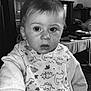 toddler, child, indoor, bib, dinosaur_pattern, hand_holding, sitting, clothing, portrait, face, expression, hair, person, table, furniture, photograph, black_and_white, nail_polish, room, background
