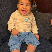 Anahat is registered to the contest to win money with this photo: baby, child, smiling, sitting, denim_pants, white_sneakers, star_pattern, beige_sweater, dinosaur_graphic, bracelets, indoor, chair, fabric_texture, cute, happy, portrait, young_child, casual_clothing, footwear, person