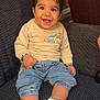 baby, child, smiling, sitting, denim_pants, white_sneakers, star_pattern, beige_sweater, dinosaur_graphic, bracelets, indoor, chair, fabric_texture, cute, happy, portrait, young_child, casual_clothing, footwear, person