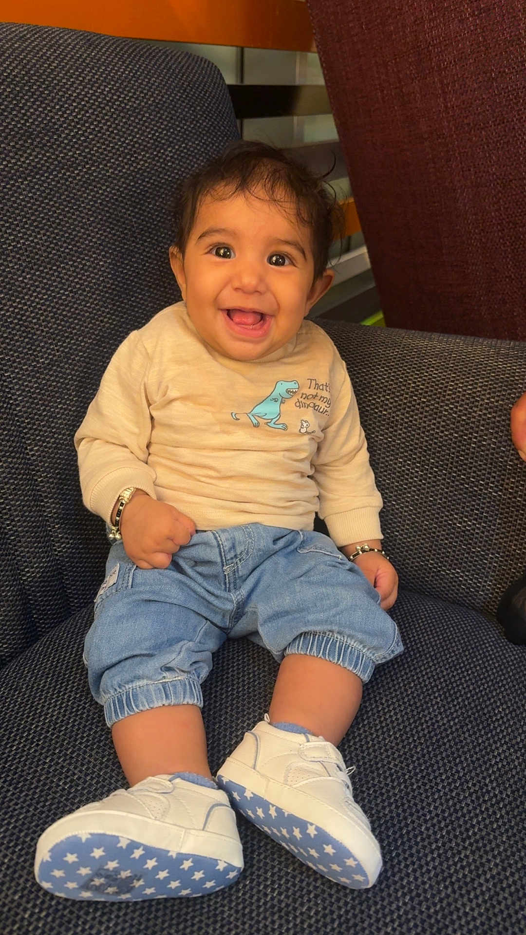 Anahat is registered to the contest to win money with this photo: baby, child, smiling, sitting, denim_pants, white_sneakers, star_pattern, beige_sweater, dinosaur_graphic, bracelets, indoor, chair, fabric_texture, cute, happy, portrait, young_child, casual_clothing, footwear, person