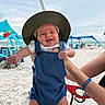 Oakley is registered to the contest to win money with this photo: baby, child, smiling, hat, blue_overalls, beach, sand, ocean, tent, summer, sun_hat, person, outdoor, happy, vacation, resort, arm, holding, sky, seaside