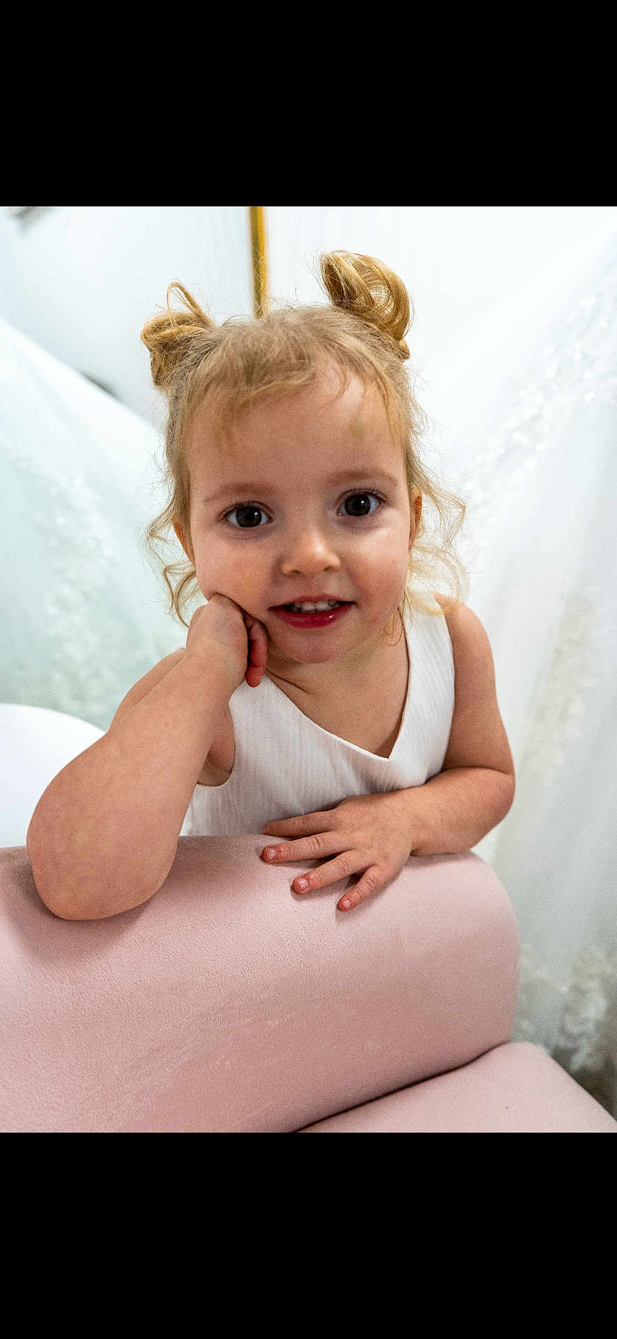Yosemite joined the competition — help win amazing prizes! child, toddler, girl, portrait, closeup, smile, hair_buns, white_dress, pink_couch, hands, arm, indoors, big_eyes, baby_teeth, curly_hair, playful, innocence, candid, furniture, young