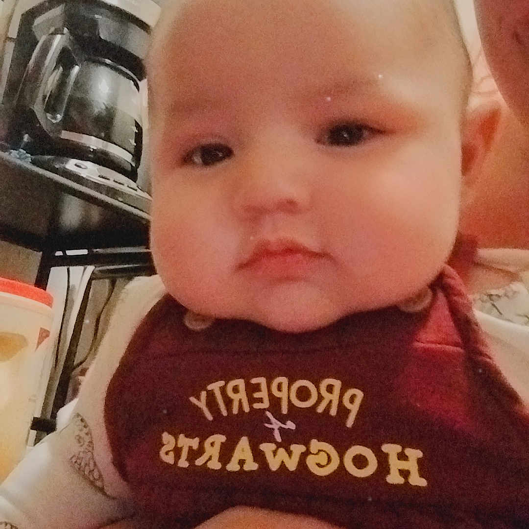 Elijah is registered to the contest to win money with this photo: appliance, baby, bib, child, closeup, clothing, coffee_maker, expressionless, face, furniture, hand, holding, indoor, infant, kitchen, maroon, person, portrait, table, text