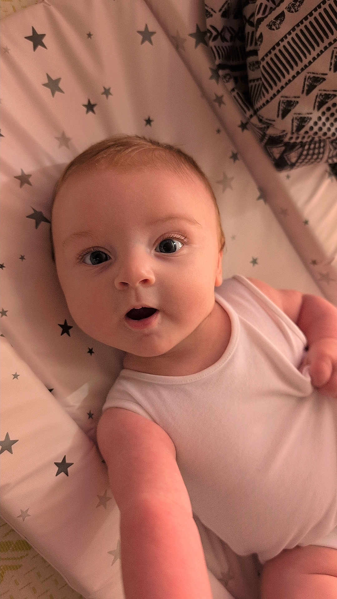 Ezra is registered to the contest to win money with this photo: baby, infant, child, onesie, pink, star_pattern, cushion, blanket, reaching, curious, face, eyes, skin, arm, indoor, closeup, person, young, expression, soft_lighting