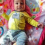 baby, child, play_mat, yellow_shirt, striped_pants, indoor, curly_hair, toy, colorful, cute, infant, person, portrait, laying_down, soft_light, happy, clothing, floor, hand, foot