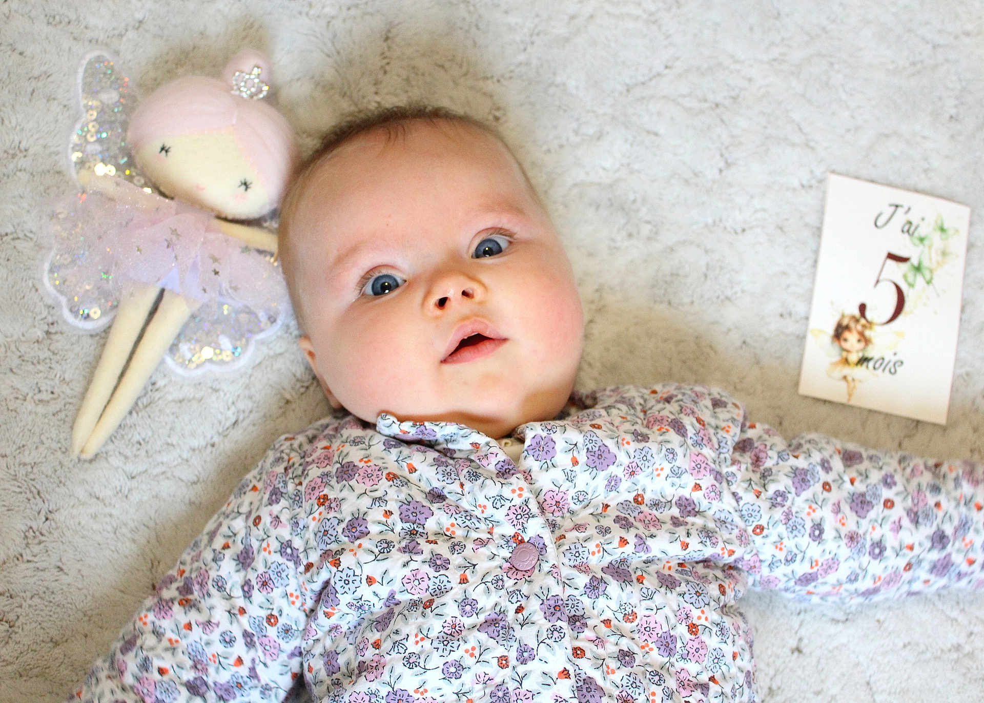 Louise a rejoint le concours — aidez-le/la à gagner de superbes lots ! baby, infant, floral_clothing, blanket, soft_texture, doll, fairy_doll, milestone_card, portrait, cute, child, blue_eyes, laying_down, indoors, closeup, face, expression, young_child, adorable, baby_girl