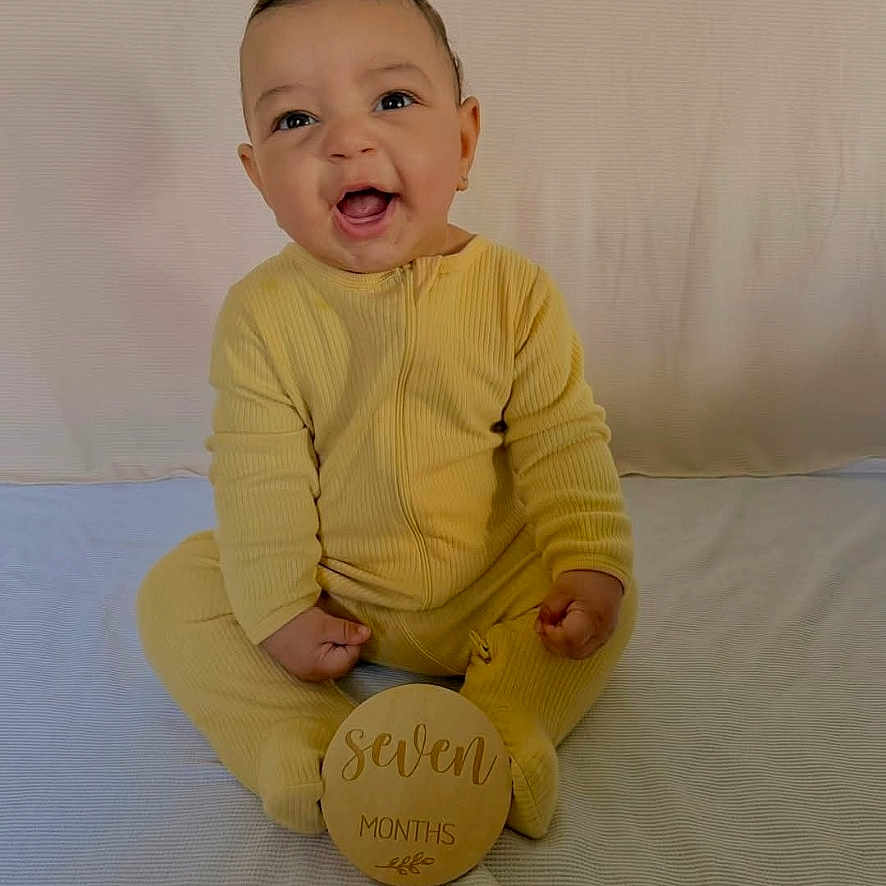 Roman is registered to the contest to win money with this photo: baby, infant, yellow_onesie, smiling, sitting, milestone, wooden_sign, seven_months, blanket, cute, portrait, child, happy, indoors, young_child, person, face, head, one_person, clothing