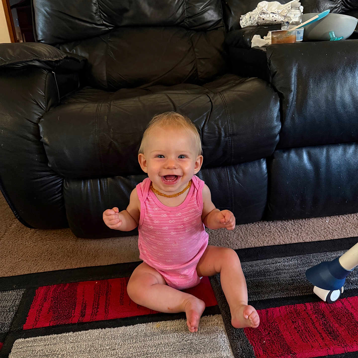 Lettie is registered to the contest to win money with this photo: baby, child, smiling, sitting, pink_onesie, carpet, black_couch, living_room, happy, indoor, person, furniture, floor, head, face, skin, hair, toy, home, cute