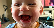Lettie joined the competition — help win amazing prizes! baby, child, smiling, laughing, face, portrait, indoors, clothing, necklace, yellow_beads, blue_shirt, happy, cute, person, head, mouth_open, teeth, living_room, television, furniture