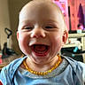 baby, child, smiling, laughing, face, portrait, indoors, clothing, necklace, yellow_beads, blue_shirt, happy, cute, person, head, mouth_open, teeth, living_room, television, furniture