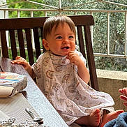Nélio participe au concours pour gagner de l'argent avec cette photo : toddler, child, smiling, table, wooden_chair, blanket, outdoor, railing, greenery, face, baby, food, spoon, foil, hand, foot, clothing, happy, dining, person