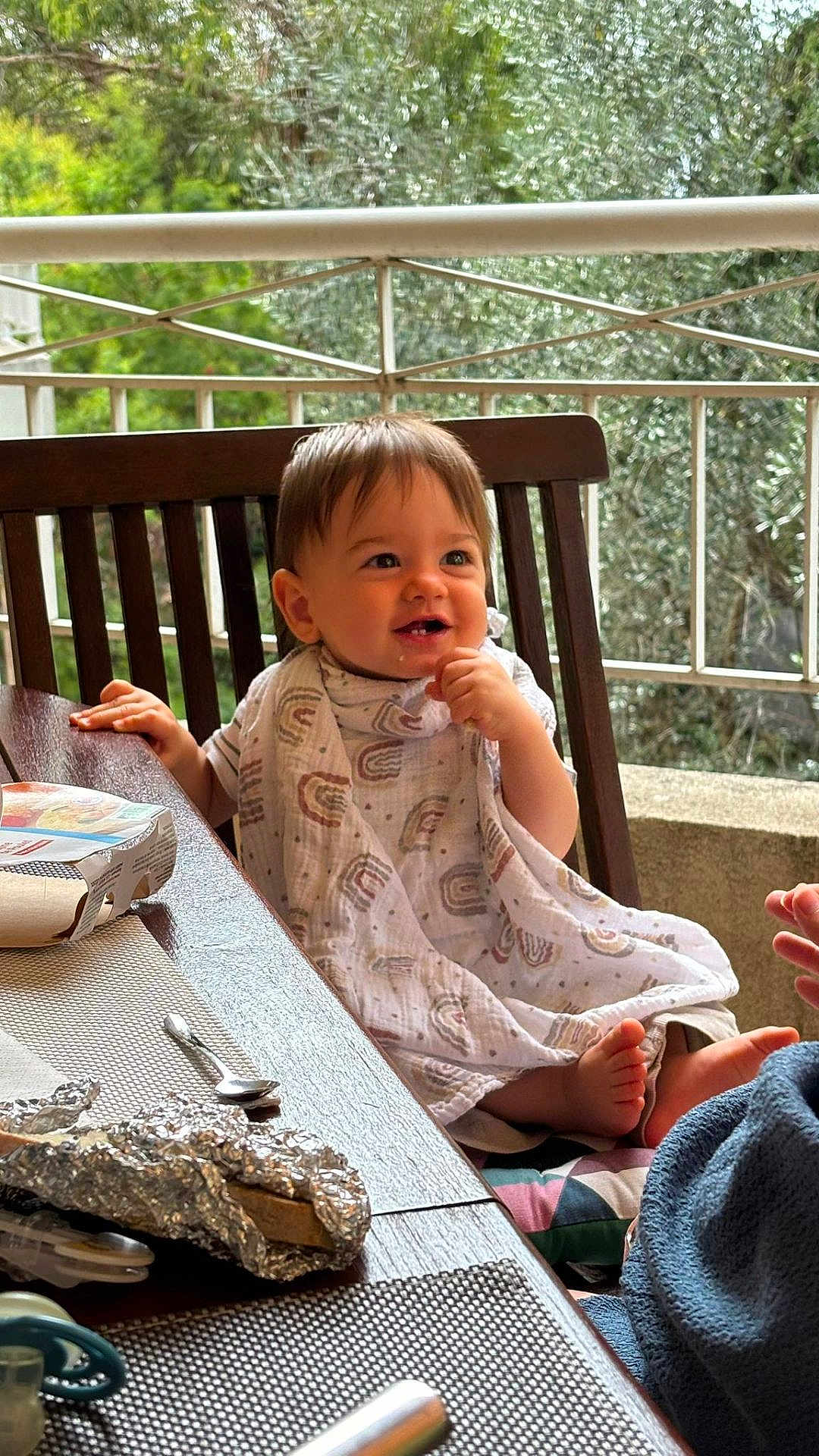 Nélio participe au concours pour gagner de l'argent avec cette photo : toddler, child, smiling, table, wooden_chair, blanket, outdoor, railing, greenery, face, baby, food, spoon, foil, hand, foot, clothing, happy, dining, person