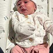 Ariella is registered to the contest to win money with this photo: baby, infant, child, white_hat, pink_pants, floral_jacket, mittens, face, tongue, cushion, patterned_background, sheep_pattern, laying_down, cute, adorable, portrait, clothing, indoor, young_child, person