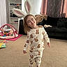 child, bunny_ears, pajamas, indoor, living_room, carpet, toys, smiling, barefoot, curtains, couch, blanket, shelf, play_mat, person, cute, happy, young_child, home, fun