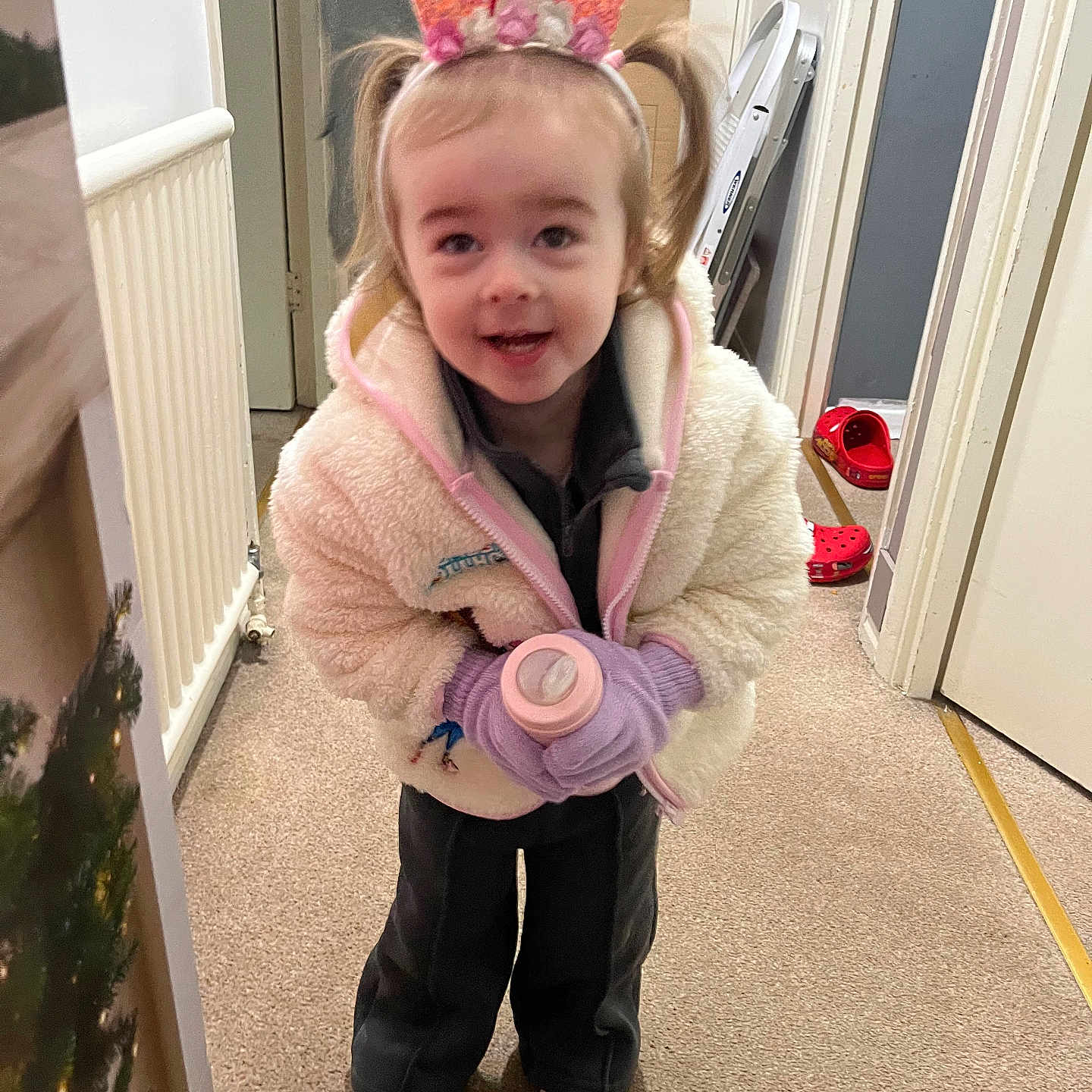 Layana is registered to the contest to win money with this photo: bottle, carpet, child, christmas_tree, clothing, crown, door, floor, footwear, gloves, happy, holiday, indoor, jacket, person, pink, shoes, smiling, toddler, toy