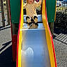 Layana is registered to the contest to win money with this photo: child, slide, playground, outdoor, yellow_dress, smiling, daylight, park, fun, happy, play, metal_slide, trees, car, fence, shadow, person, leisure, recreation, clothing