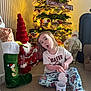 Layana is registered to the contest to win money with this photo: child, christmas_tree, holiday_decorations, pajamas, carpet, stocking, gift_box, ornaments, lights, toy, festive, indoor, holiday, sitting, expression, mug, beard, red, white, decor