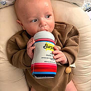 Hudson is registered to the contest to win money with this photo: baby, bottle, infant, brown_clothing, cushion, hand, face, lying_down, curious, skin, head, eyes, soft_texture, indoors, closeup, child, newborn, feeding, comfort, cute